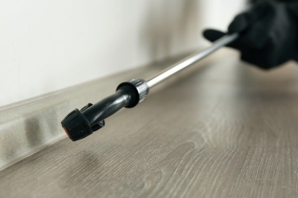 Pest control technician applying a liquid barrier treatment spray along an indoor baseboard.