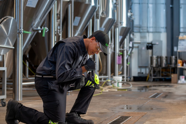 Pest control technician kneeling with a flashlight to inspect a floor drain near industrial stainless steel tanks