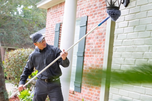 A pest control technician wearing a grey uniform, cap, and safety glasses uses a long extension pole with a grey duster attachment to sweep away cobwebs from a light fixture on an exterior brick wall.