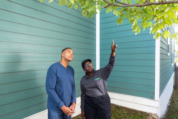 A photograph of a Terminix professional pointing up toward a tree branch while talking to a male homeowner outside a green house with wood siding.