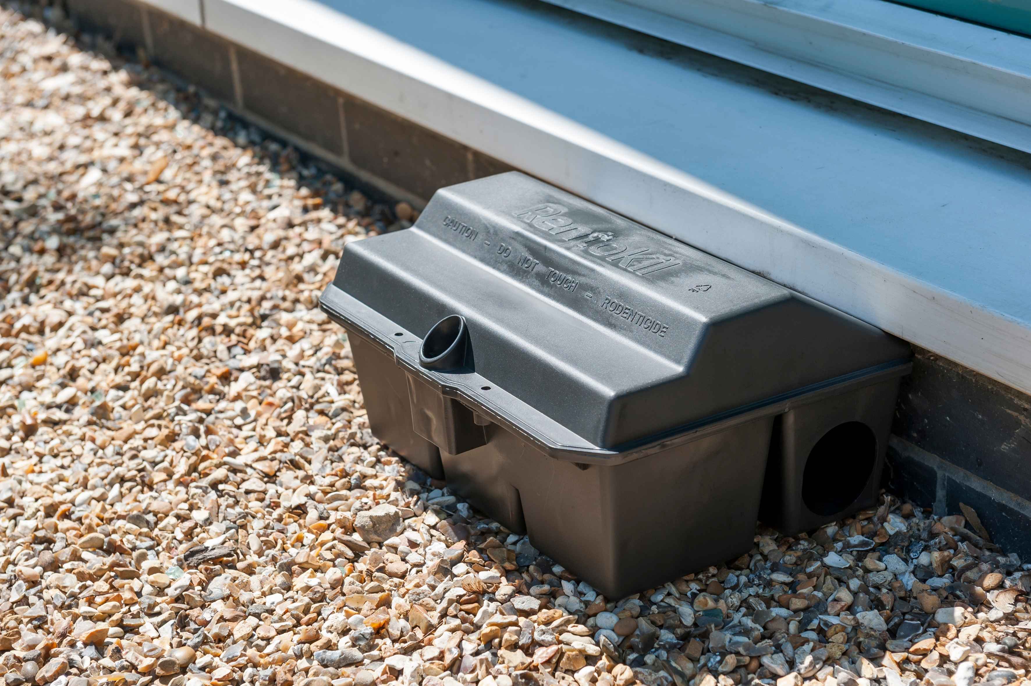 A photograph of a black plastic Rentokil rodent bait station placed on gravel along the exterior foundation wall of a building.