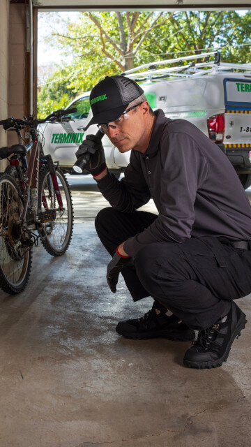 Terminix pest control technician crouching with a flashlight to inspect the interior wall of a garage, with a company truck in the background.